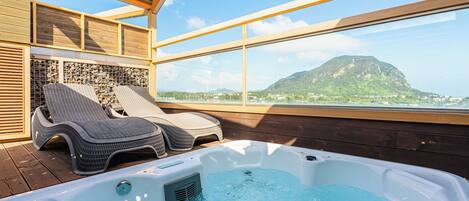 A hot tub with two lounge chairs on a wooden deck, overlooking a mountainous landscape.