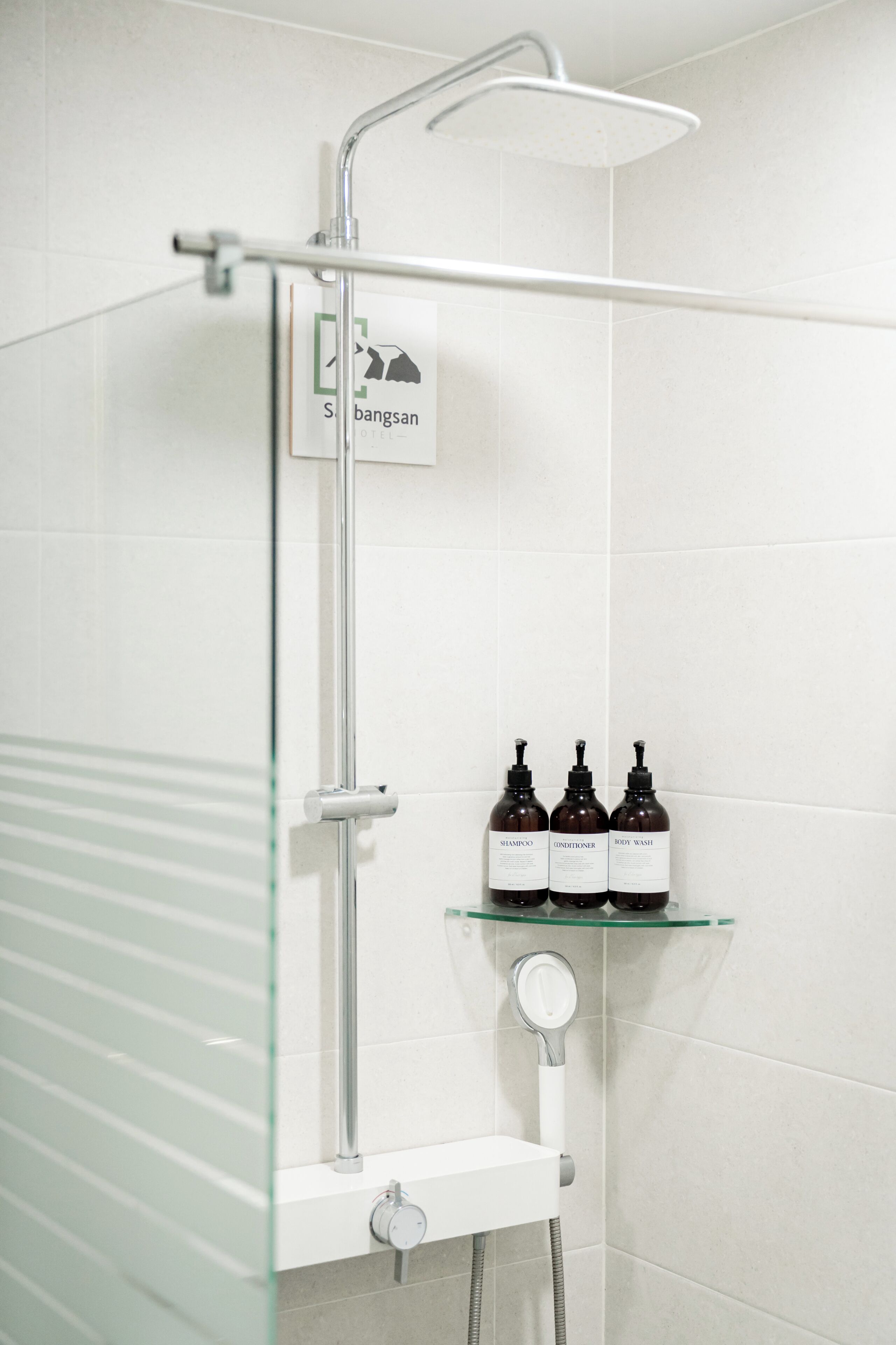 A shower area with a glass enclosure, a sign indicating a hotel, and a shelf with toiletries.