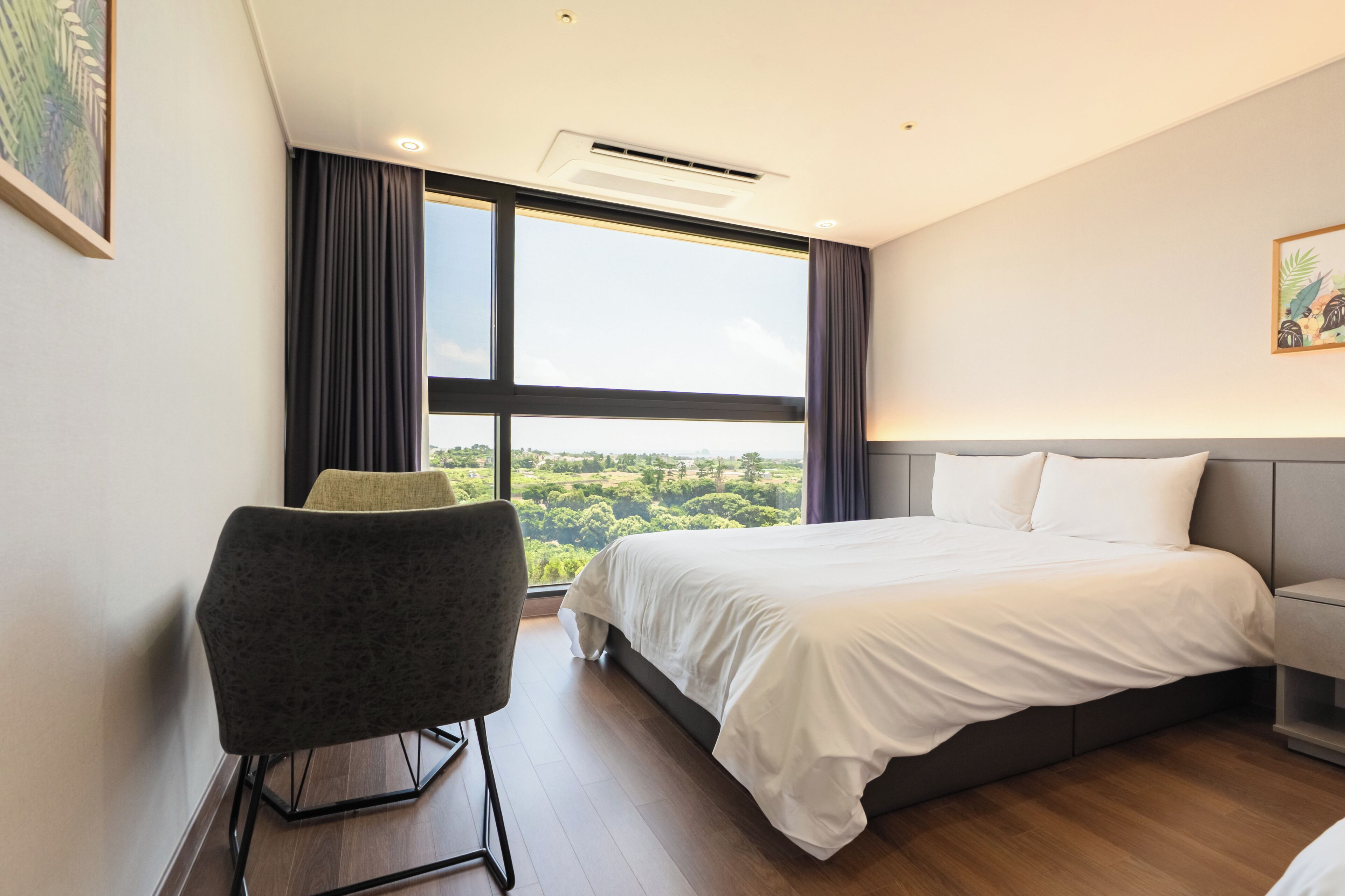 A modern hotel room with a large bed, a chair, a bedside table, and a view of the outdoors.