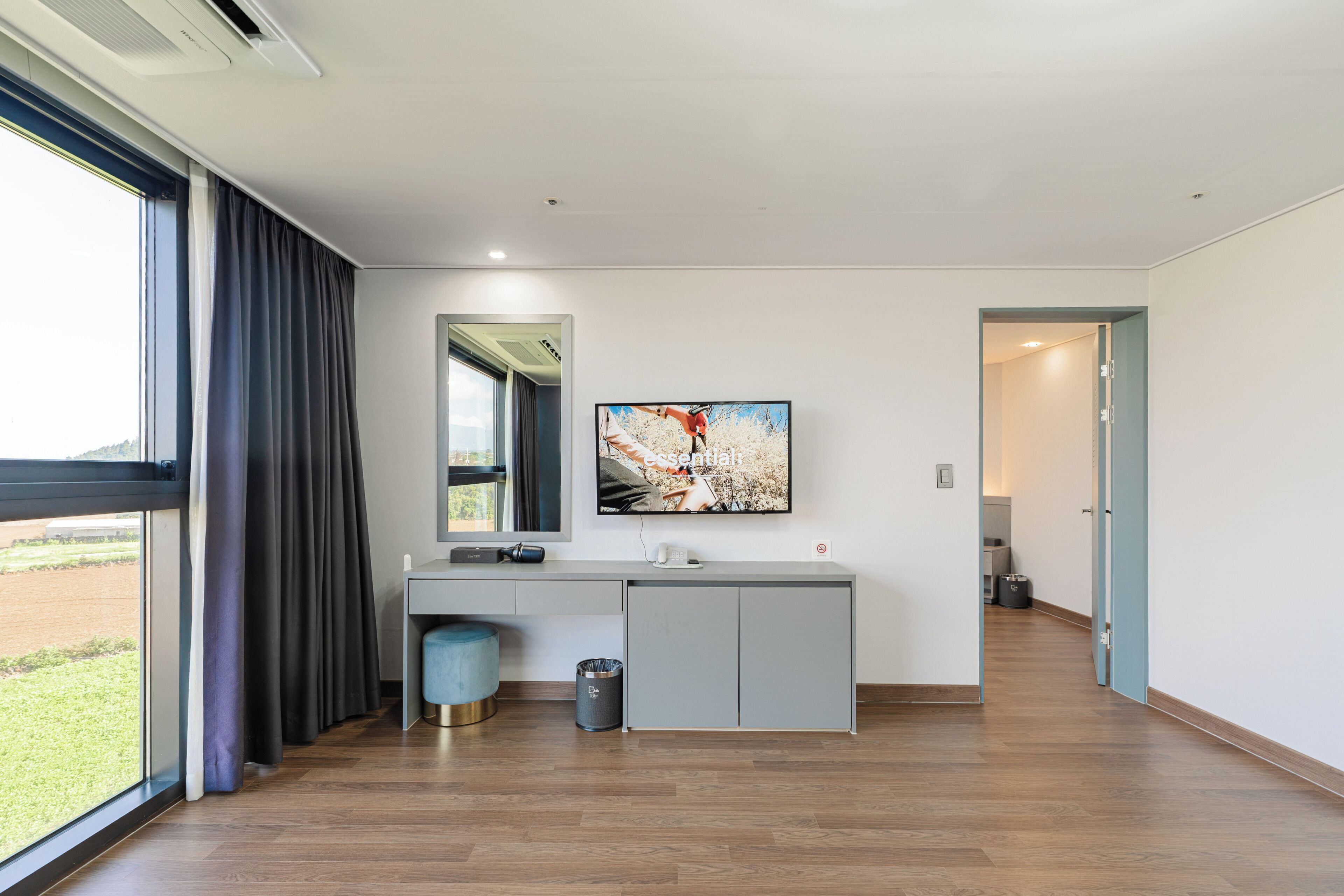 Une chambre d’hôtel moderne avec une grande fenêtre, une console grise, un téléviseur à écran plat fixé au-dessus, et un miroir sur le mur.
