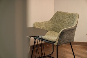 Una sola silla con asiento y respaldo de tela trenzada, junto a una mesa de madera oscura. La silla tiene un respaldo curvado y está ubicada frente a una pared lisa.