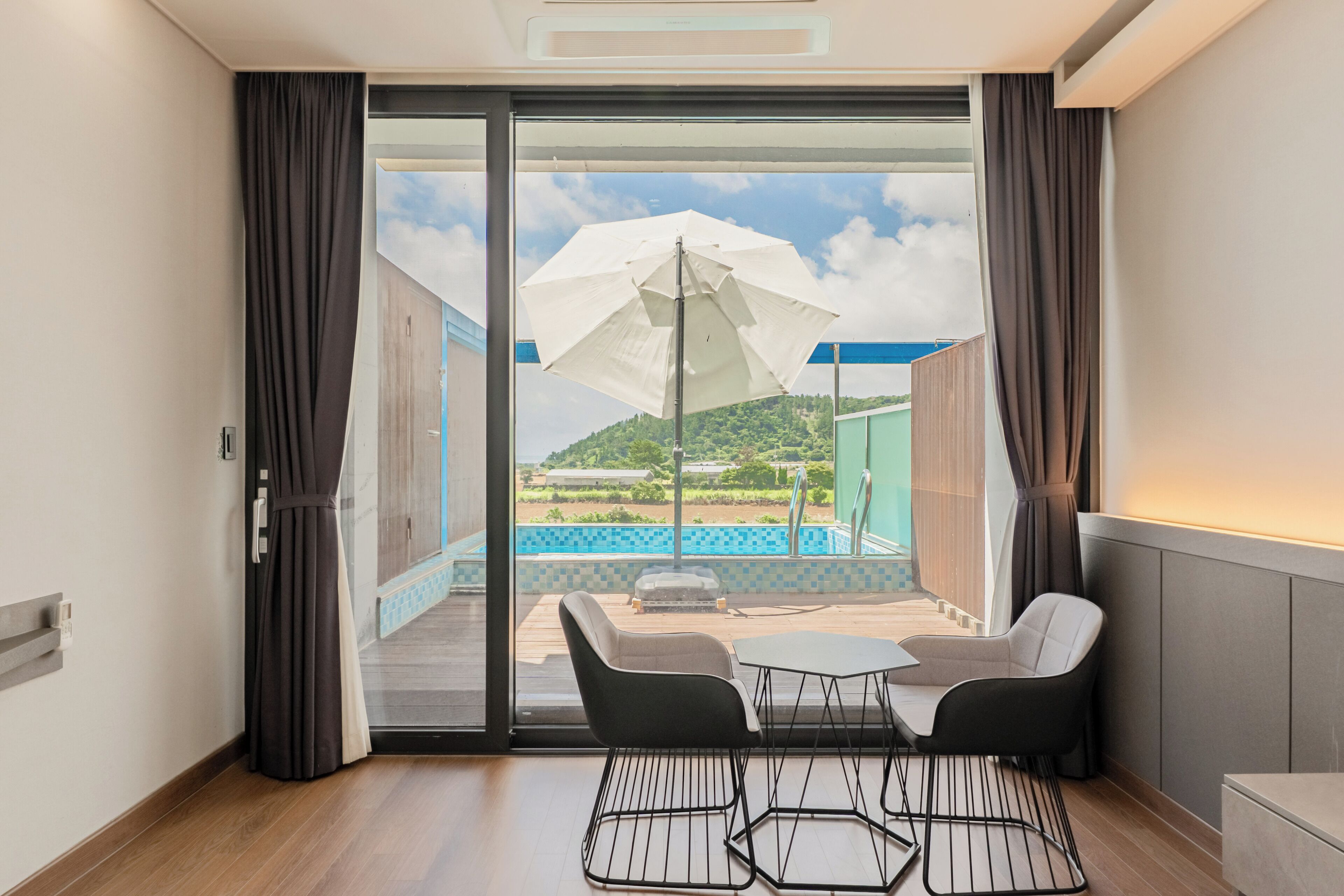 A hotel room with a sliding glass door leading to an outdoor area with a pool and umbrellas.