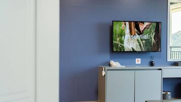 A hotel room with a flat-screen TV mounted on the wall, a desk with a telephone, and a trash can.