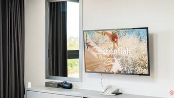 A hotel room with a flat-screen TV mounted on the wall, a bedside table with a phone and a camera, and a window with curtains.