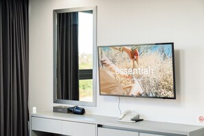 Ein Hotelzimmer mit einem an der Wand montierten Flachbildfernseher, einem Nachttisch mit Telefon und Kamera sowie einem Fenster mit Vorhängen.