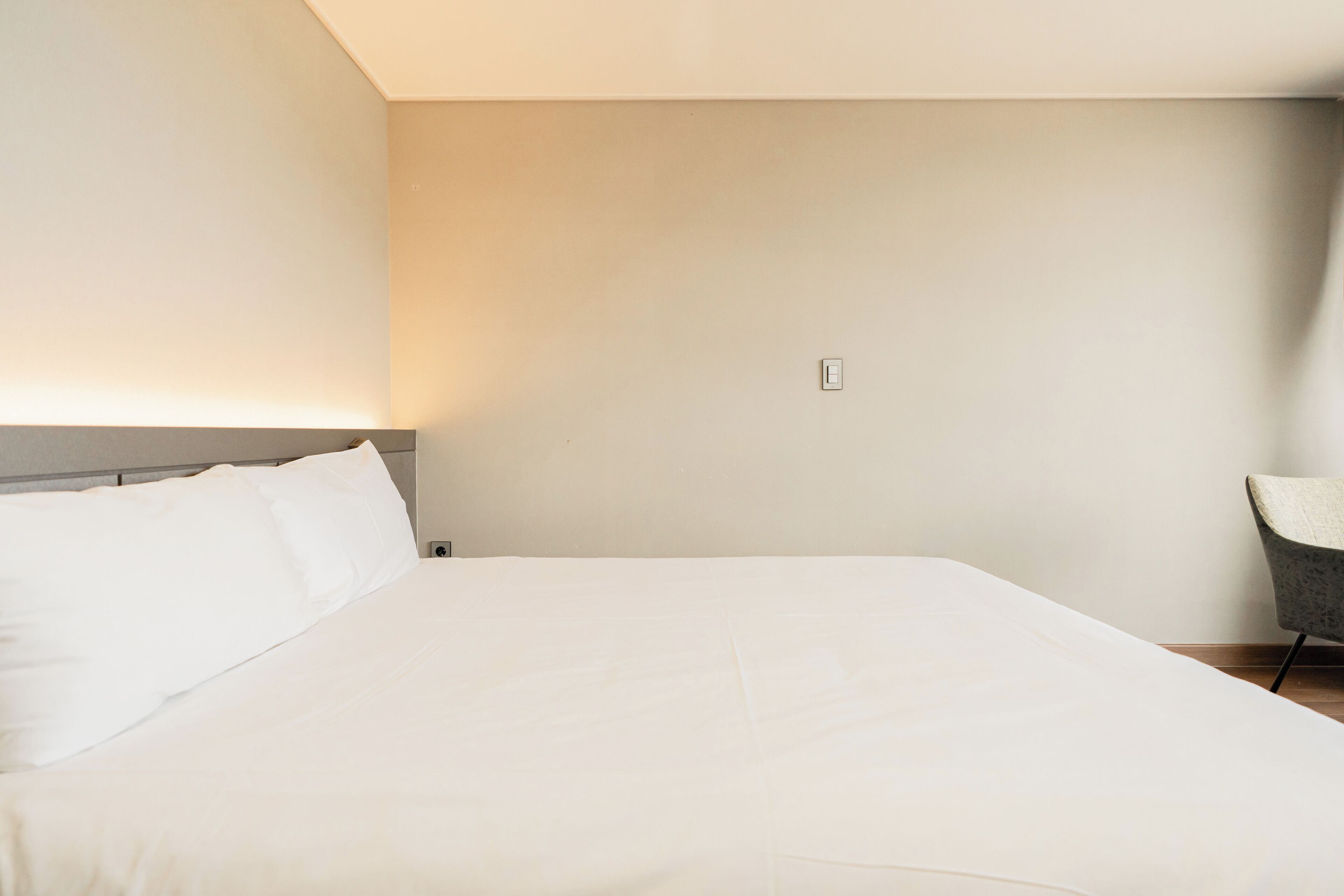Habitación de hotel minimalista con una cama grande, una silla y una lámpara de pared.