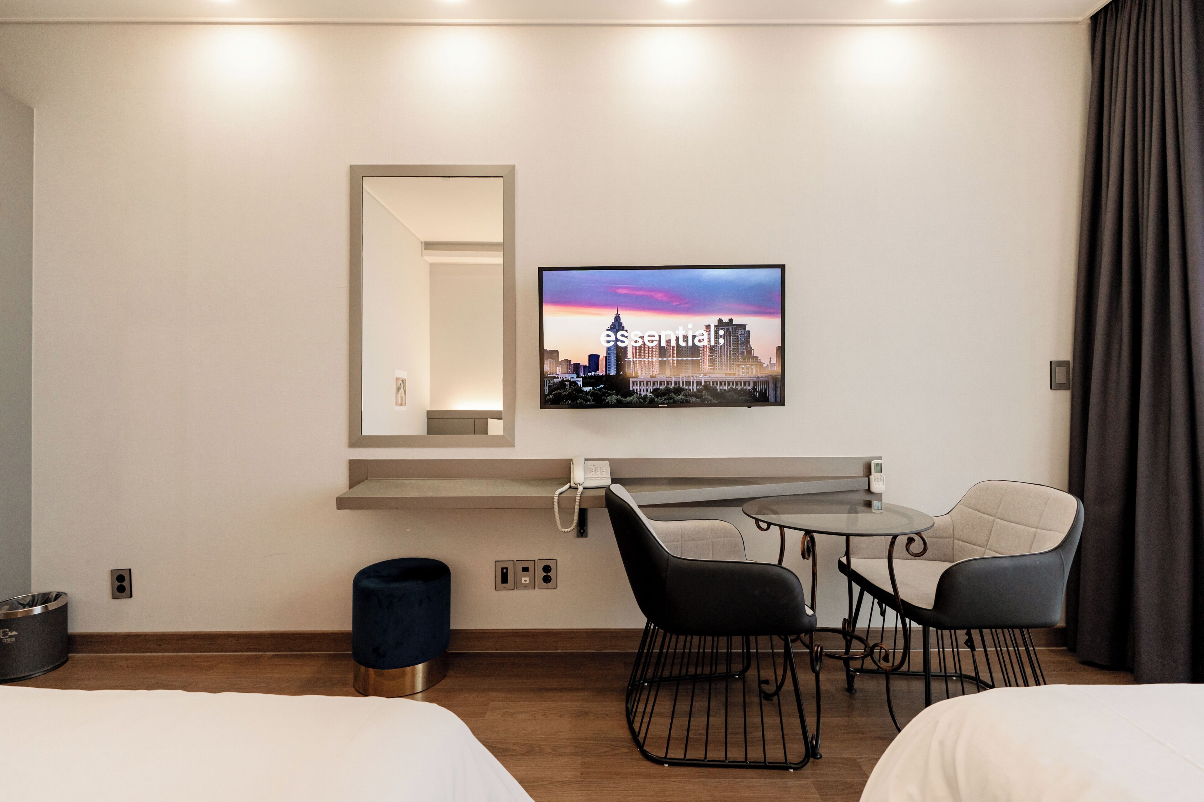 A hotel room with a bed, a desk with a chair, a small round table, a TV mounted on the wall, and a window with curtains.