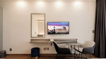 A hotel room with a bed, a desk with a chair, a small round table, a TV mounted on the wall, and a window with curtains.