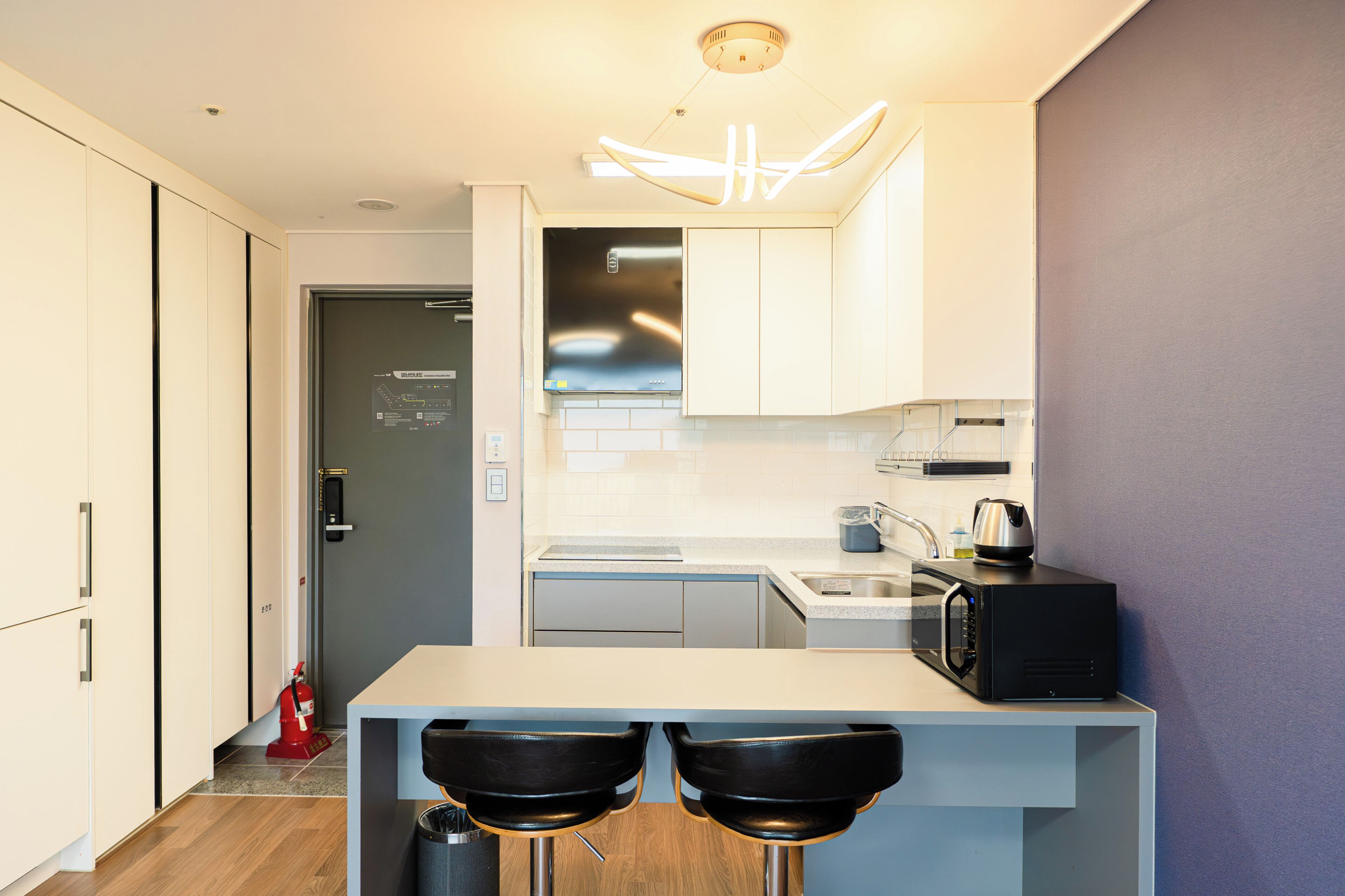 A compact kitchen with a microwave, kettle, and a small sink. There are two stools and a fire extinguisher near the door.