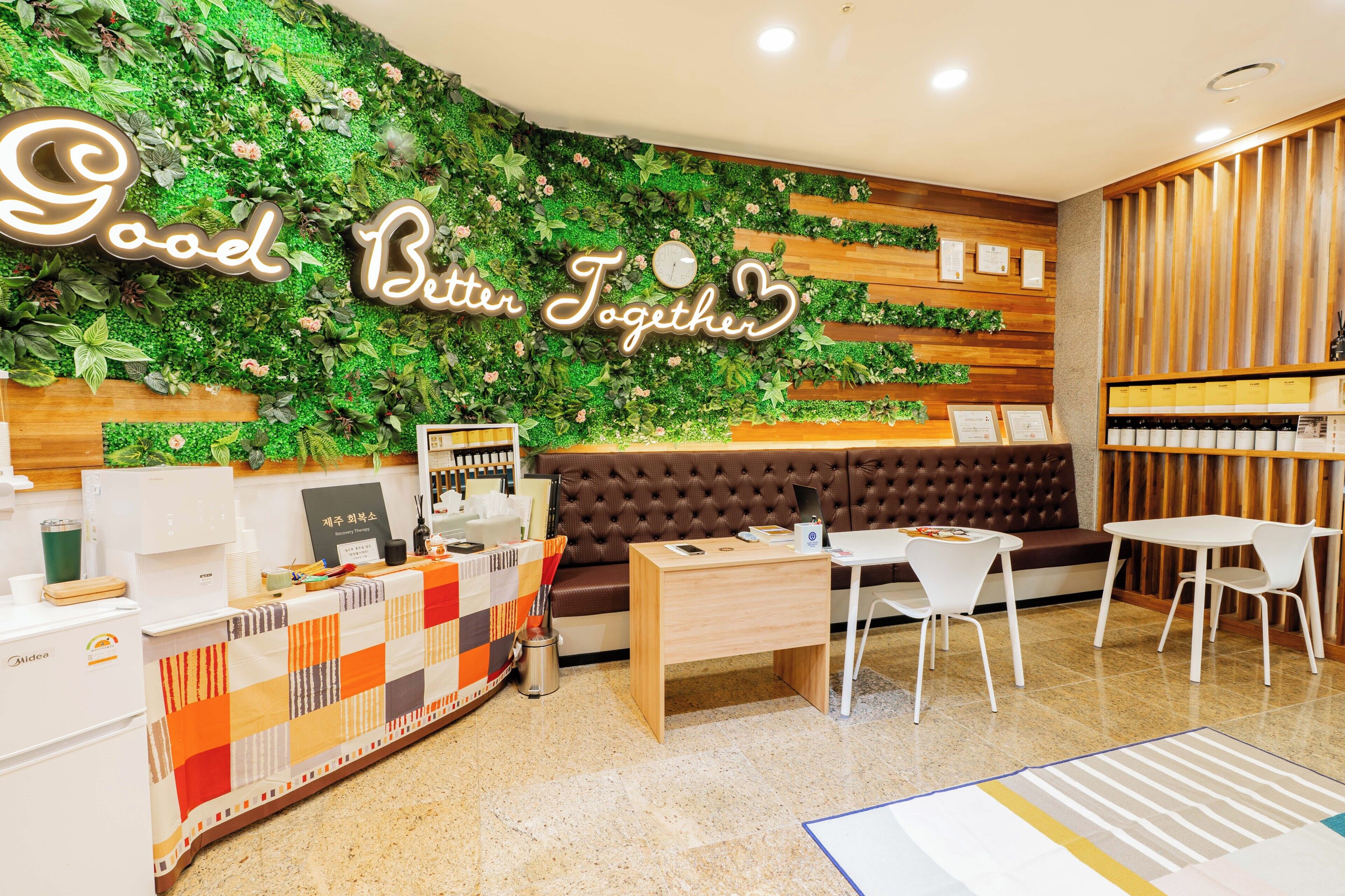 Un área de recepción con una pared verde, un letrero que dice 'Good Better Together', un mostrador de recepción, un espacio de espera con un sofá marrón y una mesita de centro.