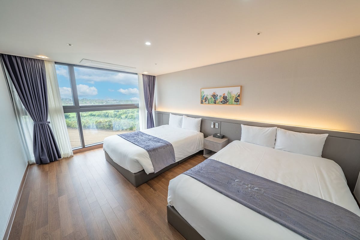 Une chambre d’hôtel moderne avec deux lits, une grande fenêtre offrant une vue panoramique et une petite table de chevet avec une lampe.