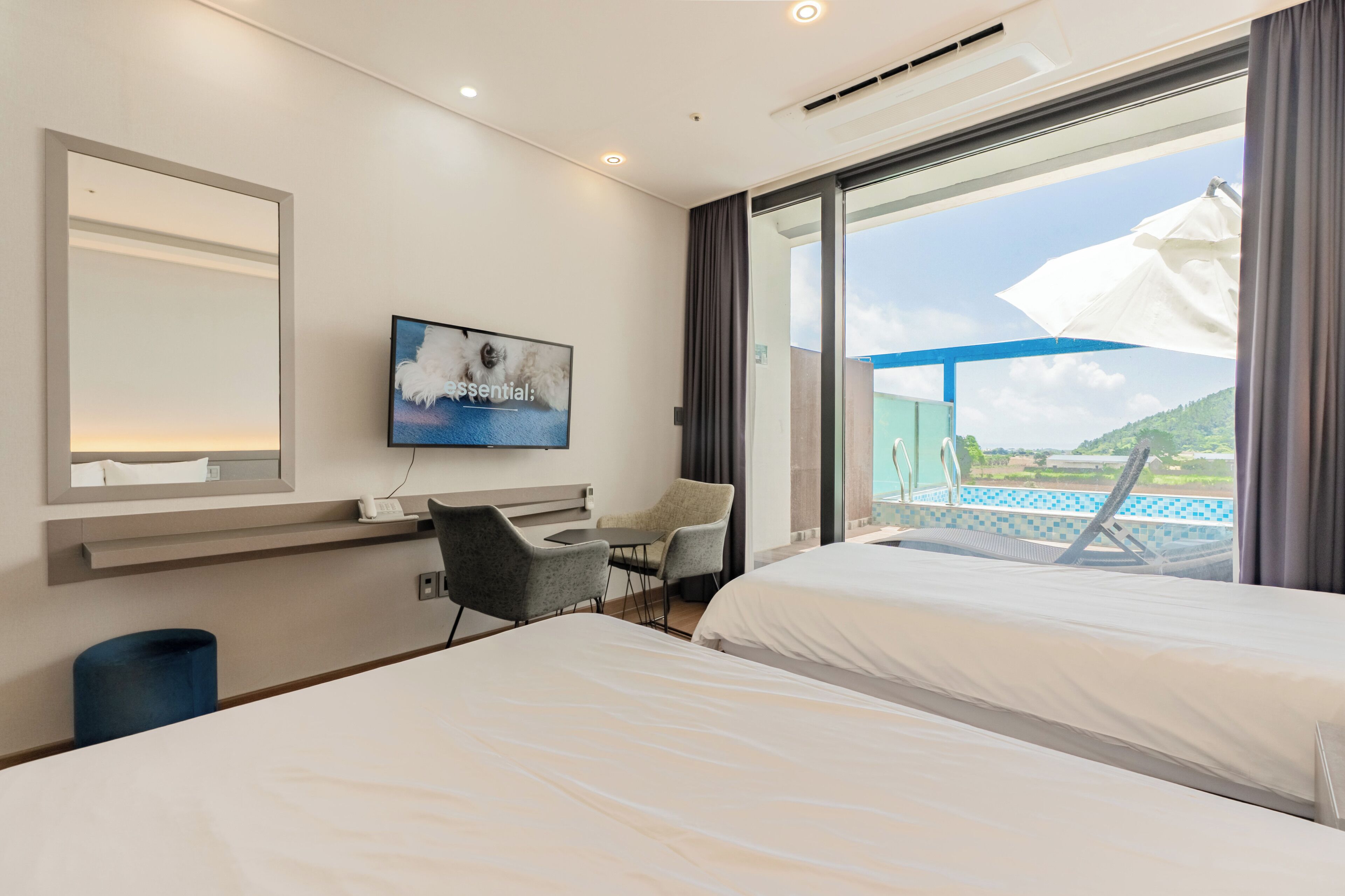 A hotel room with two beds, a desk, a chair, a TV, and a large window with a view of a pool and hills.
