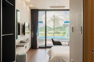 Habitación de hotel con cama, escritorio con teléfono, televisor montado en la pared y un amplio ventanal corredero que da a un balcón con piscina y mobiliario exterior.