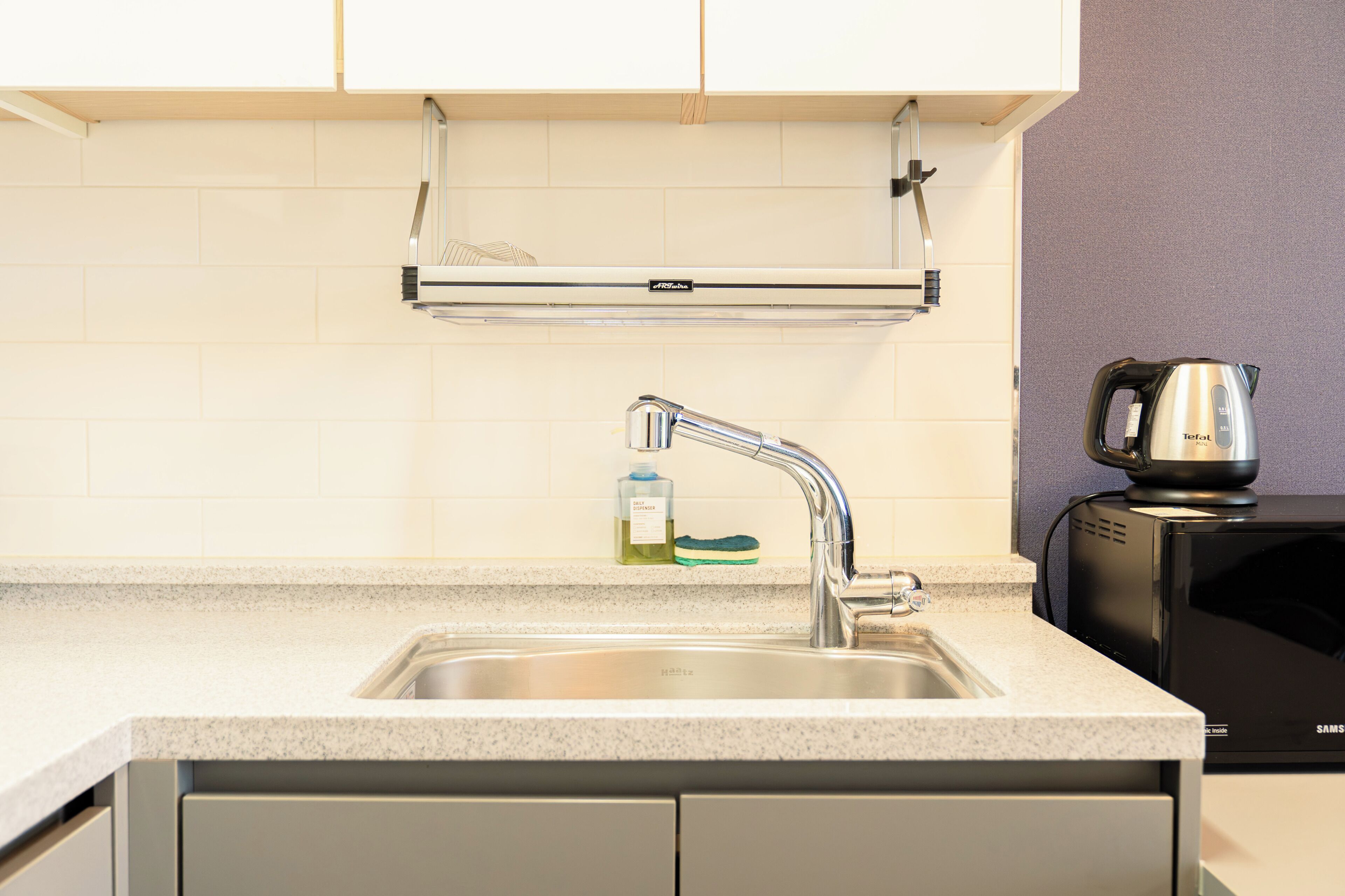 A kitchen with a stainless steel sink, a kettle, and a dish soap dispenser.