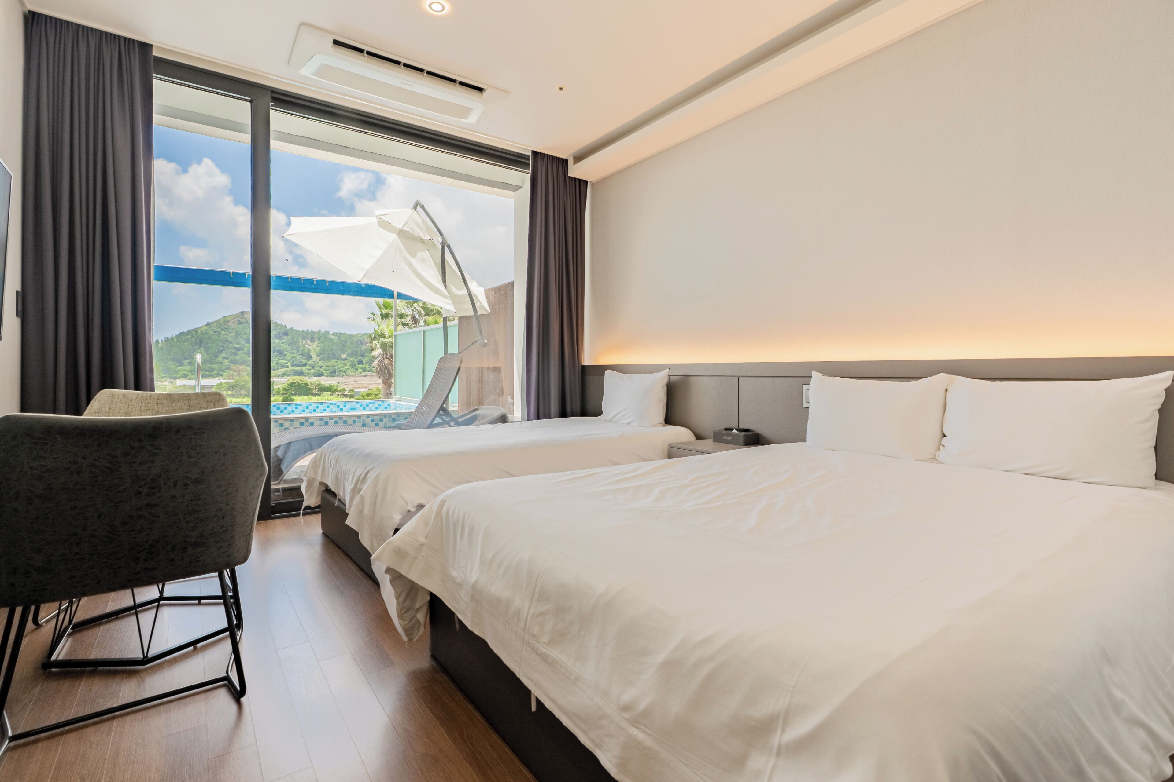 Quarto de hotel com duas camas, uma cadeira, uma escrivaninha e uma janela grande com vista para áreas verdes.