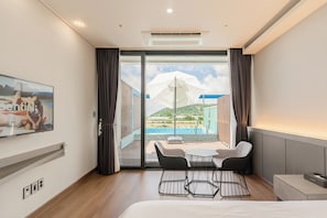 Ein Hotelzimmer mit Bett, Schreibtisch, Stuhl, kleinem Tisch, Fernseher und Blick auf Pool und Berge.