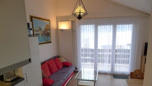 A living room with a sofa, a glass coffee table, a kitchen area with a stove, and a sliding glass door to the outside. - Elfe Apartments Studio for 2 Adults, Balcony With Lake and Mountain View (Emmetten)