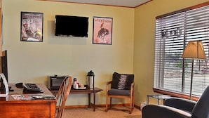 A living room with a television, two framed posters, a coffee table, a chair, and a window with blinds. - Rodeway Inn Cody (Cody)