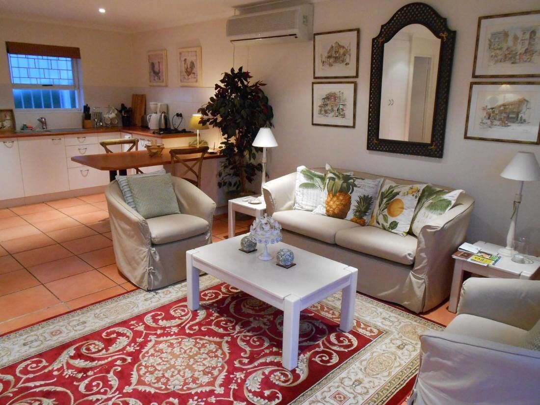 A living room with a sofa, two armchairs, a coffee table, and a dining table. There is a kitchen in the background with a window and a plant.