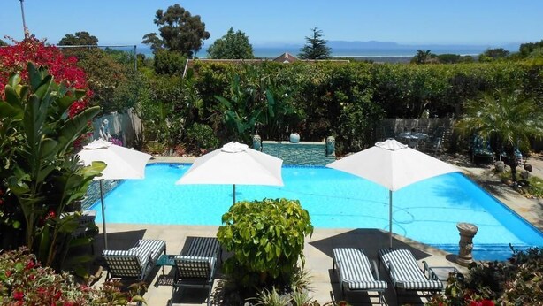 Ein Swimmingpool mit Liegestühlen und Sonnenschirmen, umgeben von viel Grün und Blick aufs Meer.