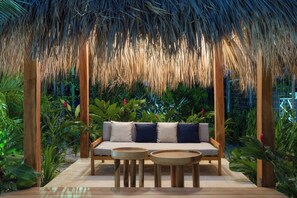 A thatched-roof outdoor seating area with a sofa and two round tables, surrounded by lush greenery.