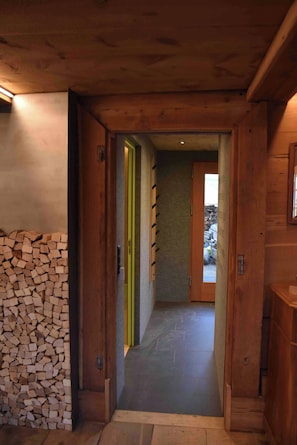 Un pasillo de madera con un montón de leña a la izquierda, una puerta verde y una vista a otra habitación.