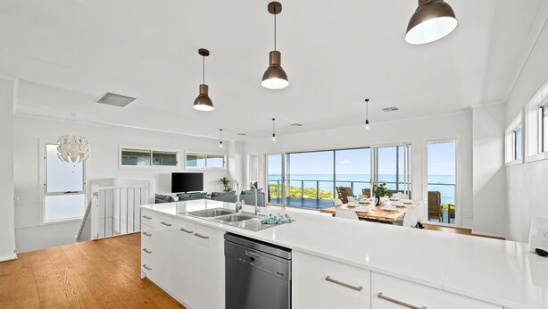 Une cuisine moderne avec des meubles blancs, un lave-vaisselle et des luminaires suspendus. De grandes fenêtres offrent une vue sur l’océan.