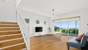 Un salon spacieux avec un escalier en bois, un téléviseur à écran plat, un canapé bleu et une grande porte-fenêtre coulissante donnant sur un balcon avec vue.