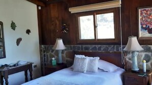 A bedroom with a bed, two bedside lamps, a nightstand, a chair, a painting, and a window with a view. - Casa Buen Aventura, Panajachel, Solola, Guatemala - Casa de Campo Familiar (Sololá)