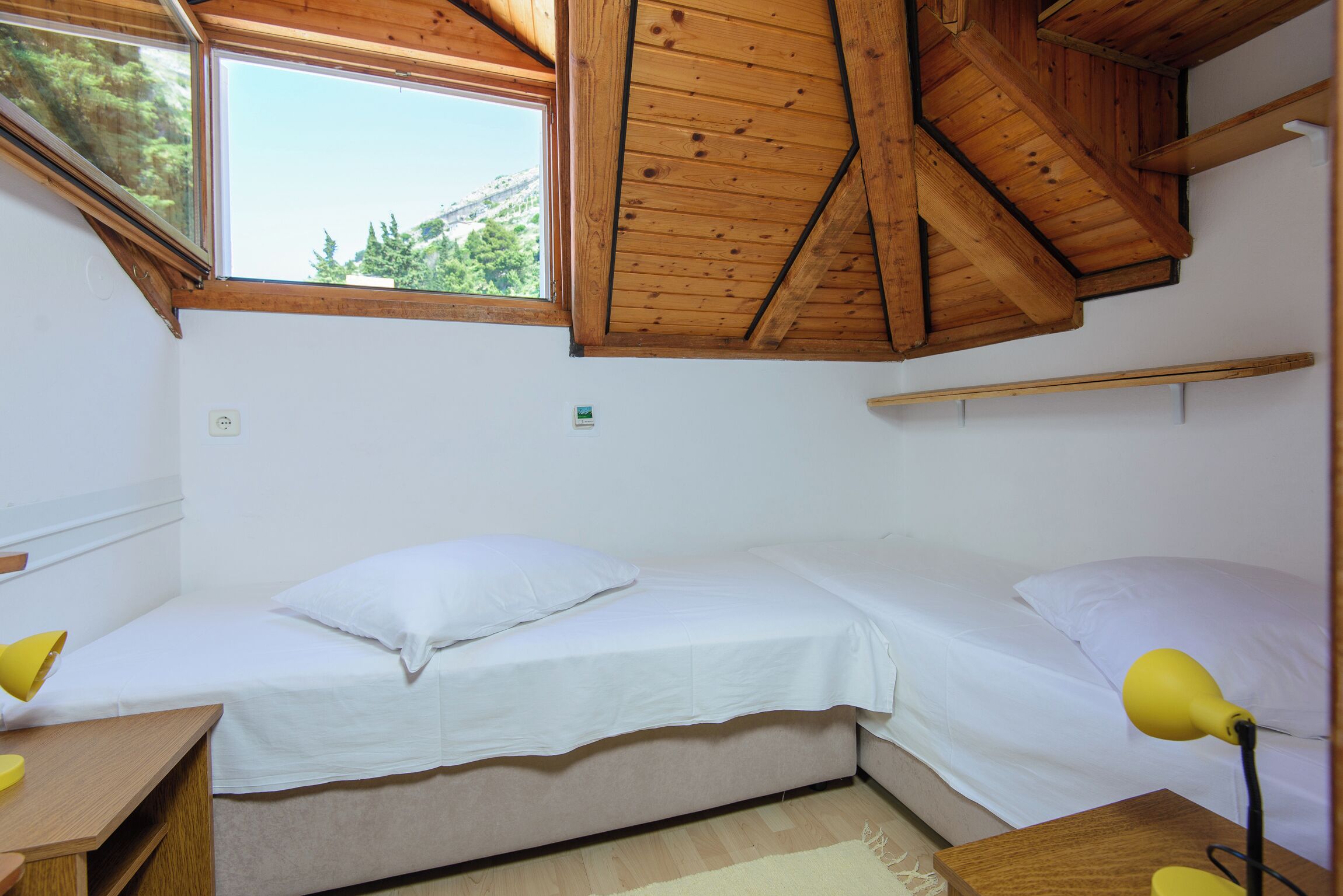 A small, single-bed room with a sloped ceiling, a window, and a desk with a lamp.