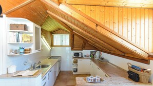Een keuken met witte kasten, een magnetron en een gootsteen. Er is een raam met zonwering en een houten plafond.