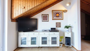 Een kamer met een houten plafond, een flatscreen televisie, een kast met lades, een klein tafeltje met een plant en decoratieve wandkunst.