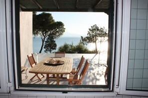 On aperçoit, à travers une fenêtre, une table et des chaises en bois sur un balcon donnant sur une étendue d’eau et des arbres.