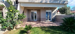 A residential building with a covered patio, two doors, and a small garden with a palm tree. - Palm Garden Apartment (Sithonia)