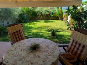 A covered patio area with a round table and two chairs, surrounded by lush greenery and a well-maintained lawn. - Palm Garden Apartment (Sithonia)