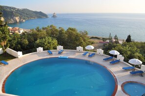 Relax by the pool with a clear view of the sea and rocky coastline, surrounded by sun loungers and umbrellas.