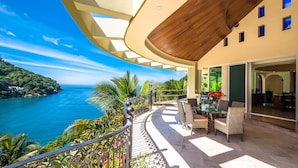 A balcony with a view of the ocean, wicker furniture, and a dining area with a table and chairs. - Beach Frontage Armonia Villa With Stunning Views (Puerto Vallarta)