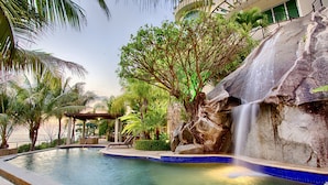 A pool with a waterfall, surrounded by palm trees and a stone wall. - Beach Frontage Armonia Villa With Stunning Views (Puerto Vallarta)