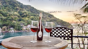 A wooden table with a bottle and two glasses of red wine, set against a scenic backdrop of a lake and forested hills.