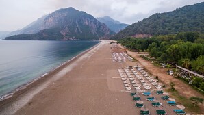A beach with rows of sun loungers and umbrellas, a clear view of the sea, and mountains in the background. - Olympos Mountain Lodge (Kemer)