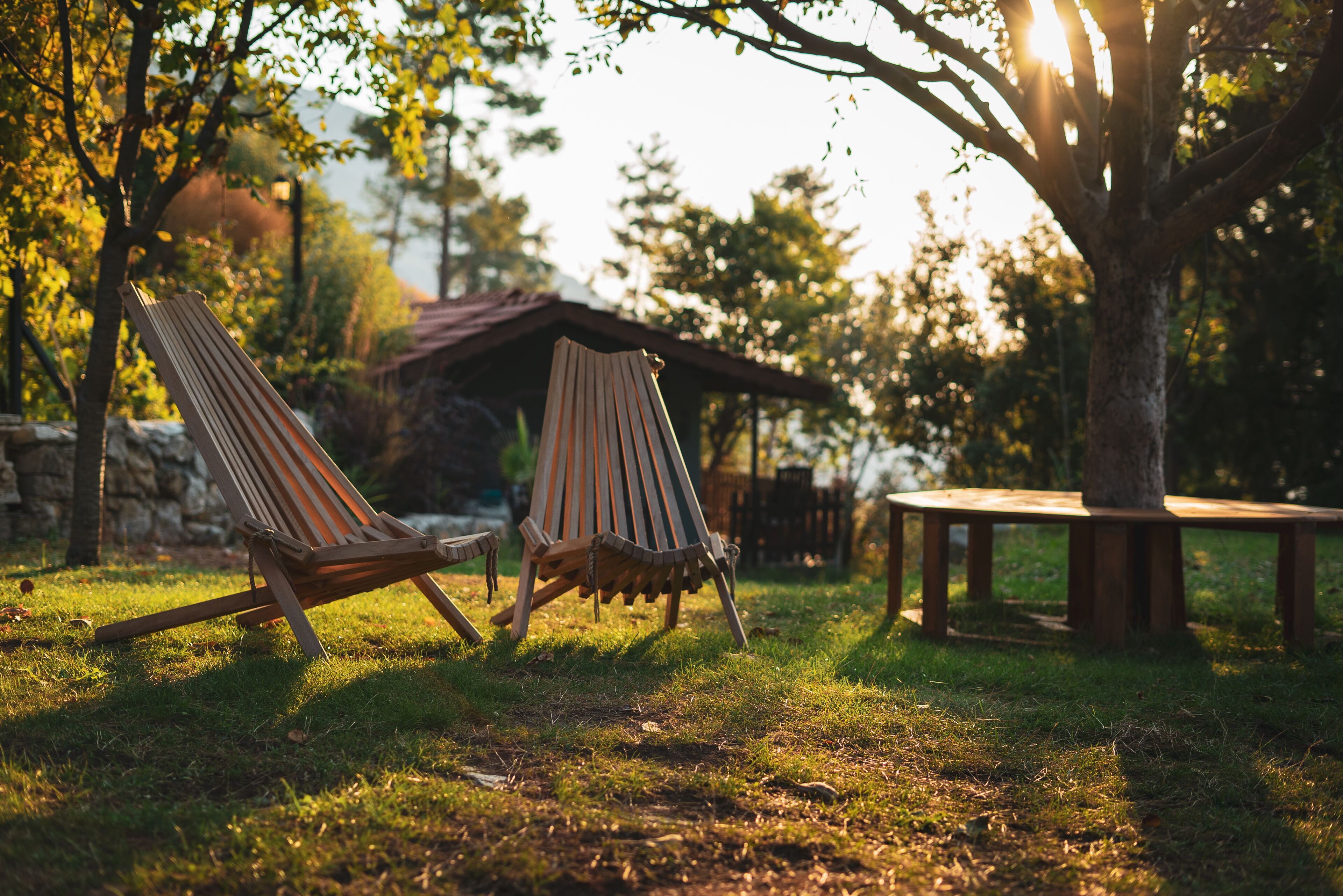 Two wooden Adirondack chairs in a grassy area with a wooden table and a stone wall in the background.