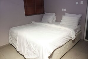 A single bed with white bedding in a room with a plain wall and a window with blinds.