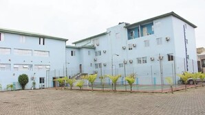 A light blue building with multiple windows and air conditioning units, surrounded by a paved area with small trees. - Room-royalrex Hotel-guest Suite (Lagos)
