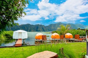 Un complexe hôtelier au bord du lac, avec des chaises longues en bois sous des parasols, une petite structure abritée et une végétation luxuriante.