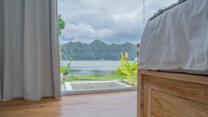 A bedroom with a wooden headboard, a bed with white bedding, and a view of a lake and mountains through a window with a curtain. - Jempana View (Kintamani)