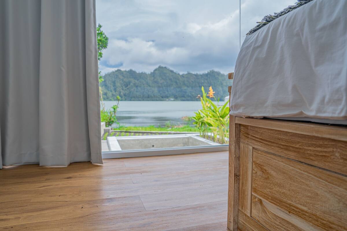 Une chambre à coucher avec une tête de lit en bois, un lit recouvert de draps blancs et une vue sur un lac et des montagnes, visible à travers une fenêtre ornée d’un rideau.