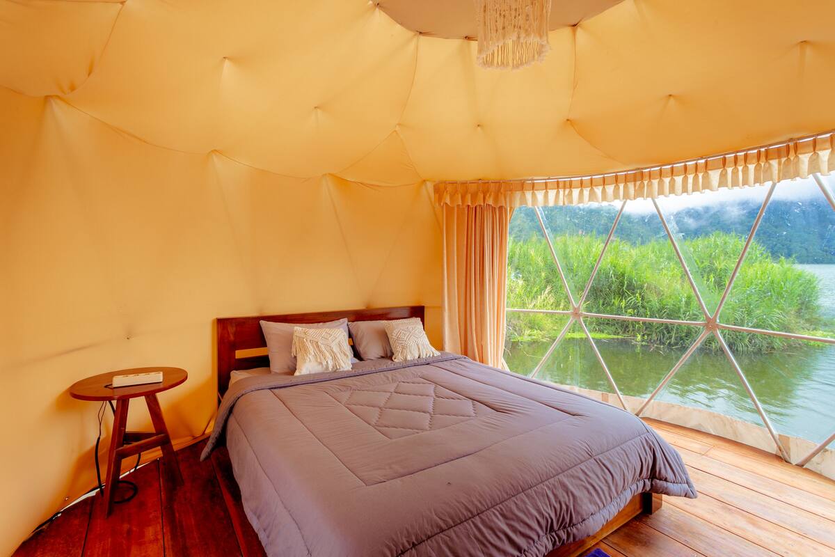 Une chambre avec un grand lit, un plancher en bois, une petite table et une fenêtre donnant sur un paysage verdoyant et un plan d’eau.