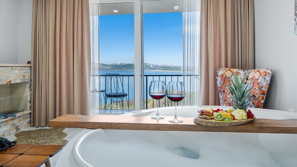A room with a large window, a wooden table with fruit and wine glasses, and a chair with a tropical print cushion.