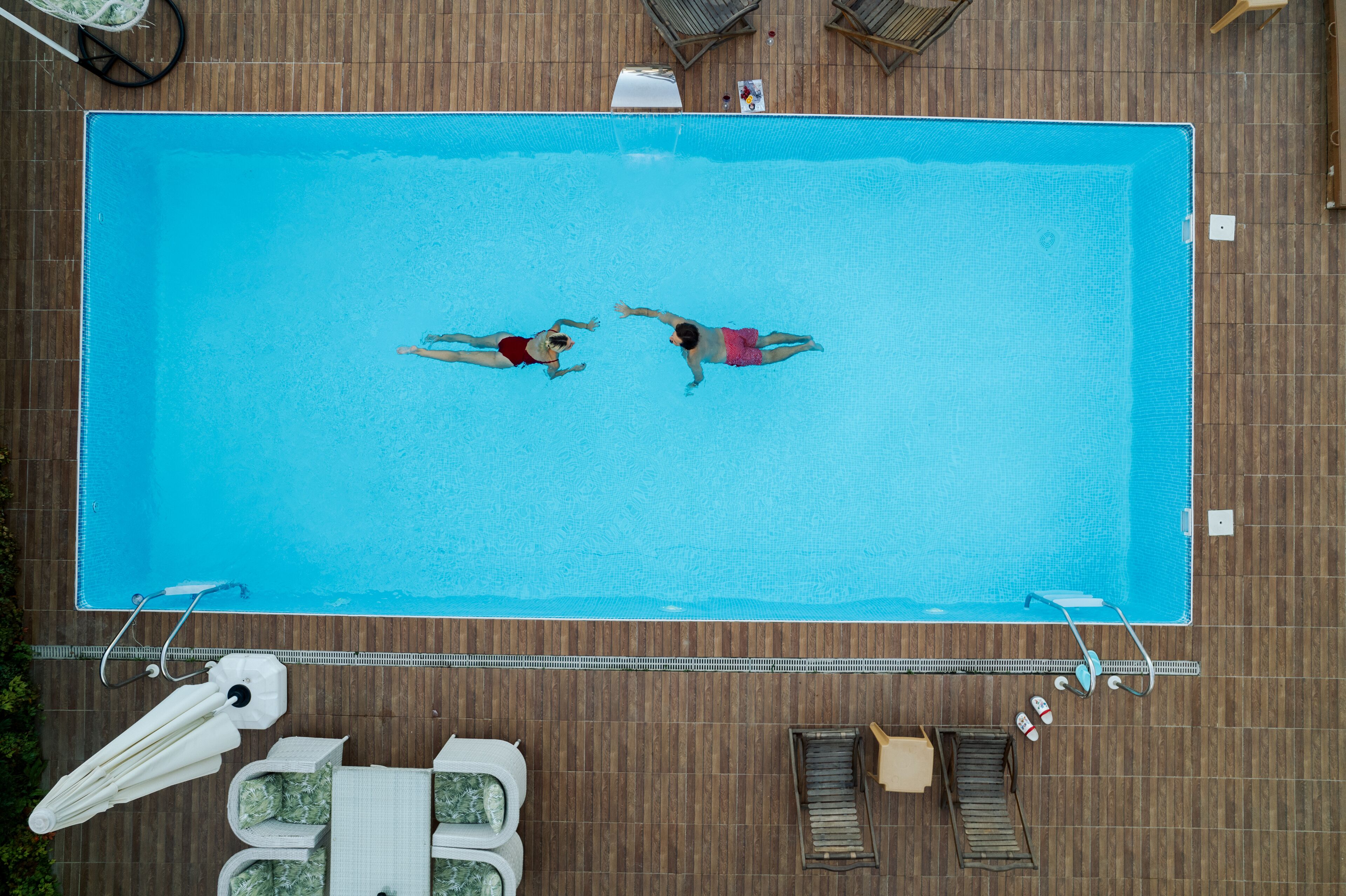Vue aérienne d’une piscine rectangulaire où nagent deux personnes, entourée d’une terrasse en bois et de mobilier de jardin.
