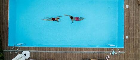 An aerial view of a rectangular swimming pool with two people swimming, surrounded by wooden deck and poolside furniture.